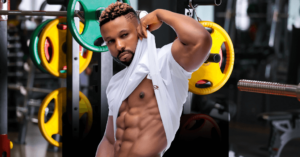 Fit man lifting his shirt to reveal well-defined abs in a gym setting, with weight plates in the background.