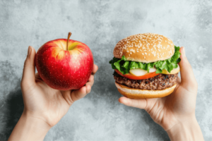 two hands holding fruit and burger, one hand holding whole food, apple while the other hand is holding processed food, burger